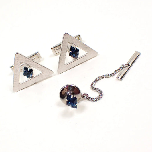 Angled view of the Mid Century vintage rhinestone men's jewelry set. The cufflinks are open triangle shaped with dark blue diamond shaped rhinestones in the middle. The metal is matte textured silver tone in color. The tie tack is a diamond shaped dark blue rhinestone stud. 