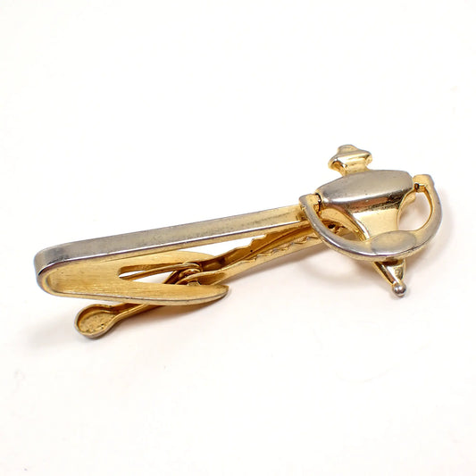 Angled view of the Hickok Mid Century vintage tie clip. It has a bar with a door knocker shape at the end. The loop on the knocker is hinged to move. The metal is gold but is showing heavy rub wear to the plating with the silver base metal showing from underneath for a two tone appearance.