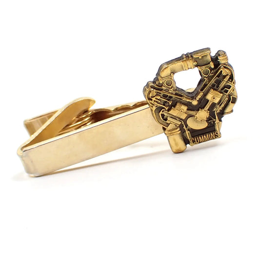 Front view of the retro vintage Cummins tie clip. The metal is gold tone in color. There is a detailed engine shape on the end with the name Cummins at the bottom.