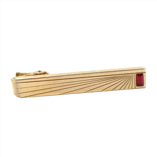 Front view of the Anson Mid Century vintage tie clip clasp. The metal is gold tone in color. There is a rectangle baguette shaped dark red rhinestone on the end. The front of the tie clip has an etched angled sun ray style design.