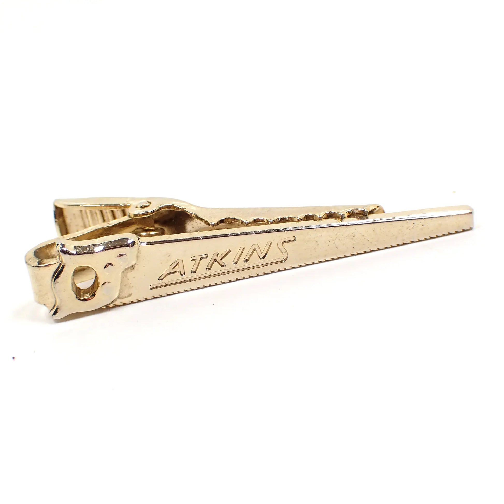 Front view of the Mid Century vintage tie clip. The metal is gold tone in color. It is shaped like a long hand saw with the company name Atkins on the front.