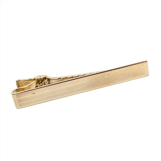 Angled view of the Mid Century vintage Anson tie clip clasp. It is gold tone in color with a long rectangular shape. There is a striped design on the majority of the front with a small rectangle at the end that is plain and shiny metal.