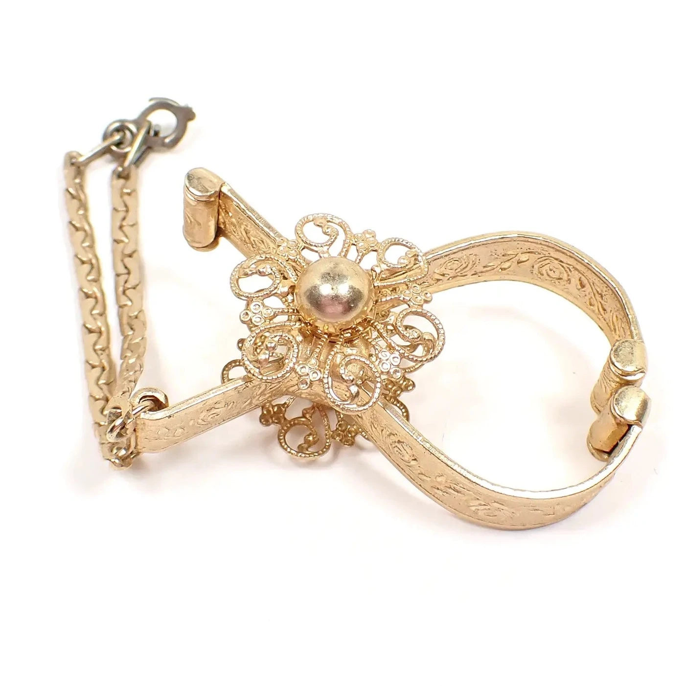 Side view of the Mid Century vintage glove holder clip. The metal is gold tone in color. It has a raised floral design and a filigree flower on each side. There is a short virola link chain at the end with a hinged clip in the middle that is darker than the rest of the piece.