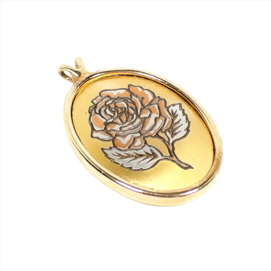 Angled view of the retro vintage Reed and Barton pendant. It is oval in shape. There is an etched rose flower design on the front with copper and silver color accents. The rest of the pendant is gold tone in color. There is a double opening V shaped bail at the top. There is no chain.