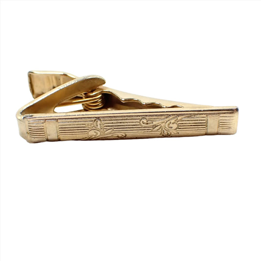Front view of the Mid Century vintage tie clip clasp. The metal is gold tone in color. The front has a striped design with two areas of scroll like leaves.
