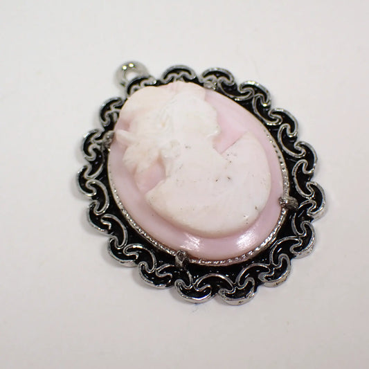 Retro vintage cameo pendant. The setting has an antiqued silver color with fancy edge. The oval cameo is made of light pink and white glass with the pink primarily as the background. It is of a woman's head bust. There are tiny dark spots seen around the neck and bottom area in the photo.
