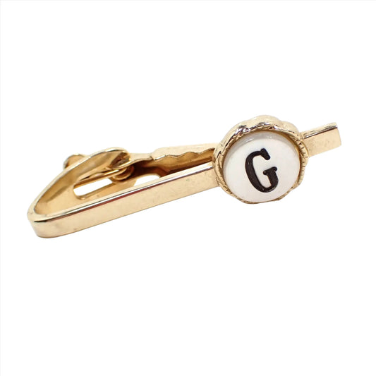 Angled view of the retro vintage initial tie clip clasp. The metal is gold tone in color. There is a round area on the end with a pearly white flat mother of pearl shell cab. The cab has an engraved and black painted letter G in the middle.