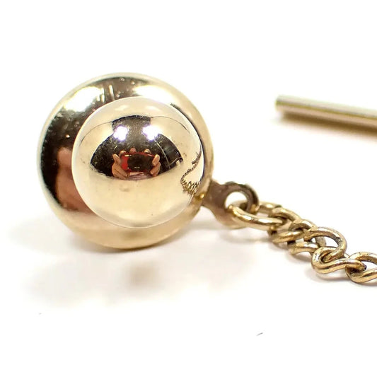 Close up of the Mid Century vintage tie tack. The metal is gold tone in color. The tie tack itself is round ball shaped and shiny. You can see my reflection taking the photo in it. The clutch and part of the clutch chain can be seen in the photo.