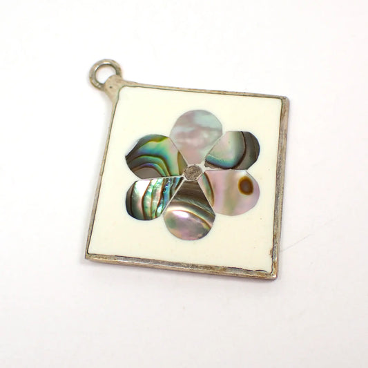 Front view of the retro vintage Alpaca Mexico pendant. It is diamond shaped with silver tone color metal. The front is white enameled with inlaid abalone shell in a flower design with rounded petals.