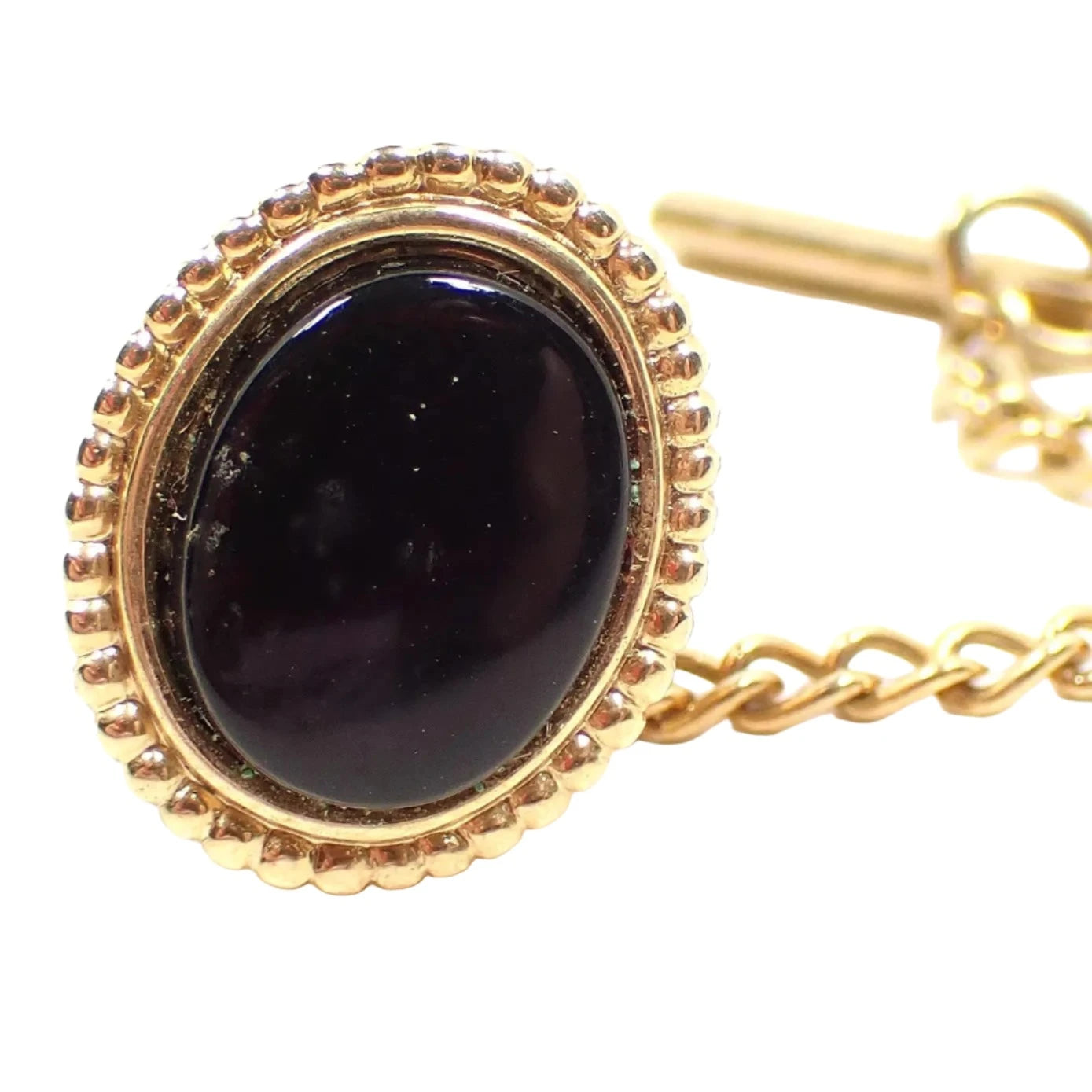 Front view of the Retro vintage Avon tie tack. The metal is gold tone in color and is oval in shape. The middle of the oval has a black glass cab. The outer edge has round dots all the way around it. From the back there is a chain with a bar at the end.