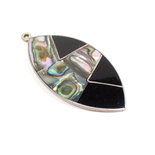 Angled view of the retro vintage Alpaca pendant. It is marquis shaped with silver tone color metal. There are areas that are black enameled on the right and bottom of the pendant and the rest has three pieces of inlaid abalone shell that is multi color iridescent.