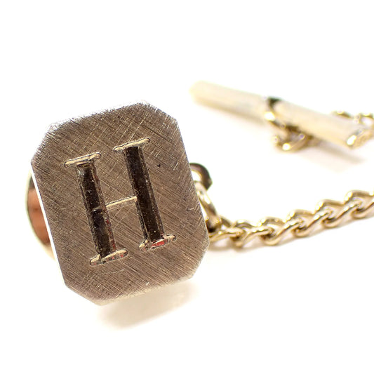Front view of the Mid Century vintage initial tie tack. It is octagon shaped with brushed matte gold tone color metal. There is a letter H engraved on the front.