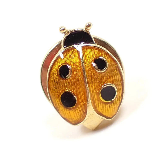 Front view of the retro vintage ladybug tie tack. The metal is gold tone in color. The lady bug has an orange yellow enameled body and black enameled spots and head. The clutch on the back is round and is the type without a chain.