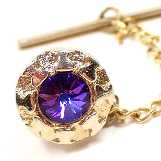 Front view of the Mid Century vintage rivoli rhinestone tie tack. The metal is gold tone in color. It has a round shape with textured metal cut out areas around the edge. The rhinestone comes to a point in the middle and flashes shades of blue and purple as the light hits it.
