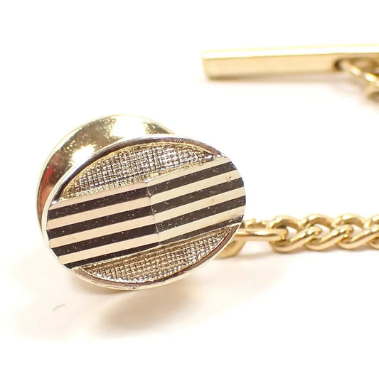Enlarged front view of the Mid Century Modernist vintage tie tack. The metal is gold tone in color. It is shaped like an oval and has textured matte background with an angled etched line design in the middle.