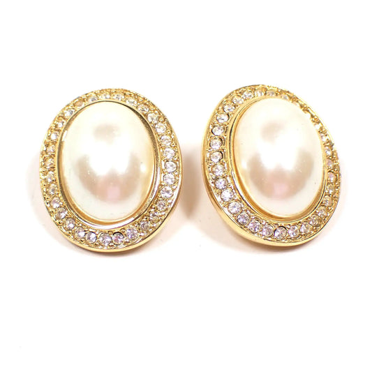 Front view of the retro vintage Marvella clip on earrings. They are oval in shape with gold tone color metal. The fronts have domed plastic faux pearl cabs in a light off white color. They are surrounded by a row of small clear rhinestones.