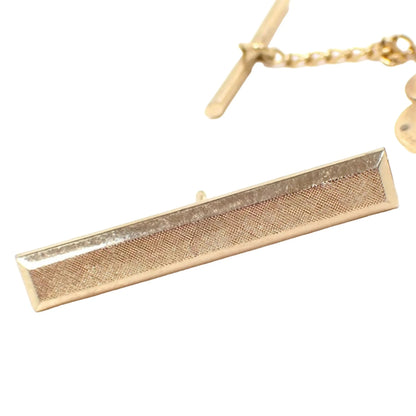 Front view of the Swank Mid Century bar tie tack. It has a long rectangle bar shape with matte textured front and shiny metal edges. The metal is gold tone in color. A clutch back with chain can be seen in the background.