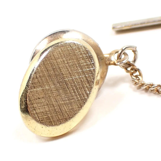 Front view of the Mid Century vintage tie tack. The metal is gold tone in color and the front has a brushed textured matte appearance. It is oval shaped and the clutch back with chain can be seen in the photo.