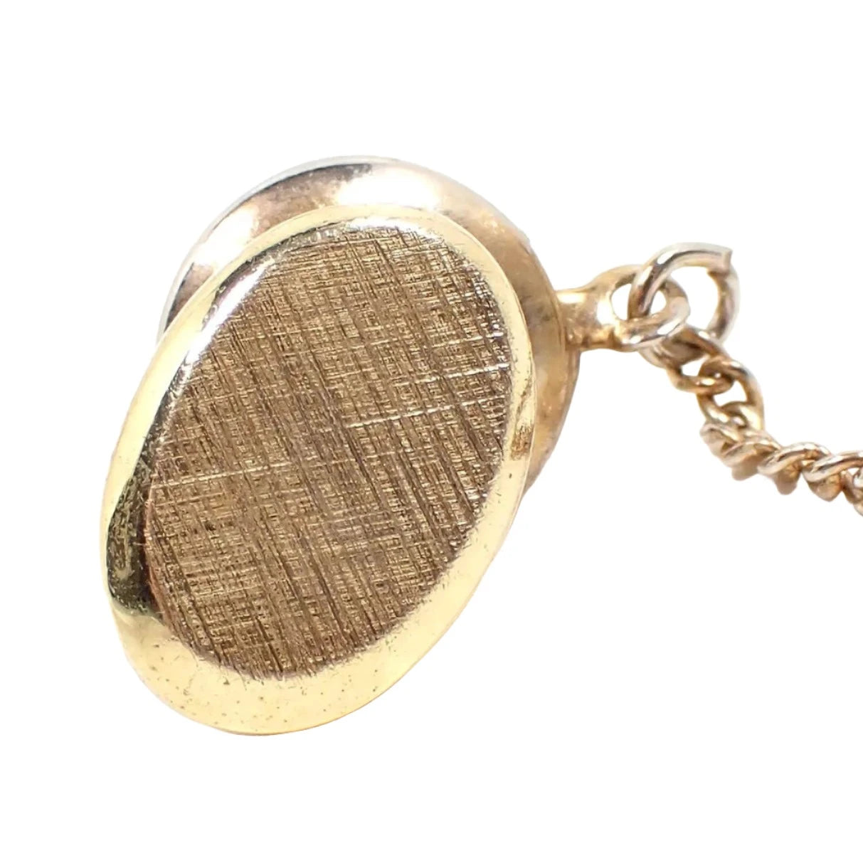 Front view of the Mid Century vintage tie tack. The metal is gold tone in color and the front has a brushed textured matte appearance. It is oval shaped and the clutch back with chain can be seen in the photo.