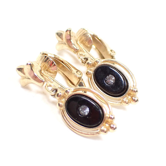 Angled view of the retro vintage Avon clip on earrings. The metal is gold tone in color. The drops are oval shaped with black glass cabs that have a tiny clear rhinestone in the middle of them.