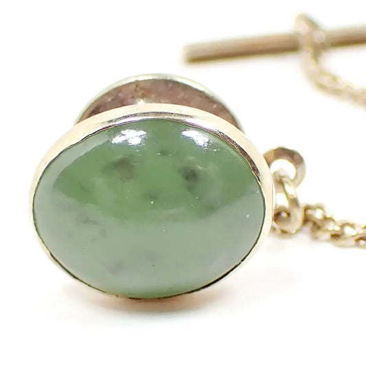 Front view of the retro vintage gemstone tie tack. It is oval shaped with a Chrysoprase gemstone cab. The cab is a semi translucent light green color with flecks of darker green within. The metal is gold tone in color. The clutch back and chain shows in the background.