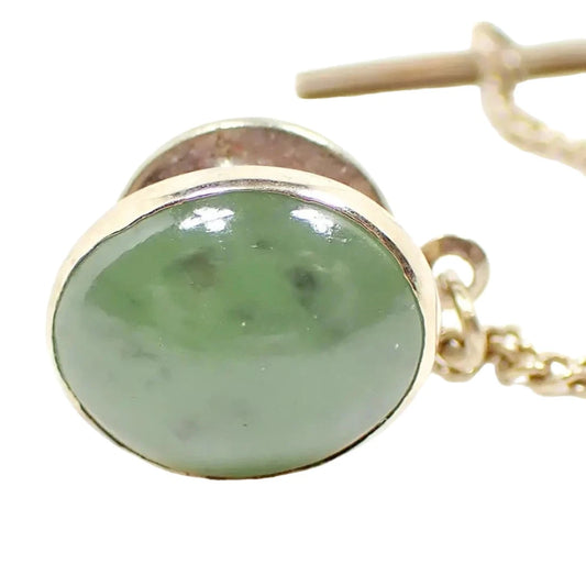 Front view of the retro vintage gemstone tie tack. It is oval shaped with a Chrysoprase gemstone cab. The cab is a semi translucent light green color with flecks of darker green within. The metal is gold tone in color. The clutch back and chain shows in the background.