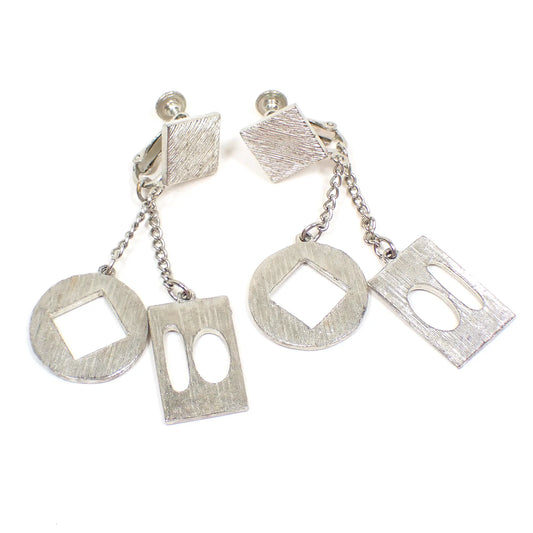 Angled view of the Mid Century vintage Atomic geometric dangle clip on earrings. The tops are diamond shaped and there are circle and rectangle drops with oval and square cut out designs on them. The metal is brush textured silver tone in color.