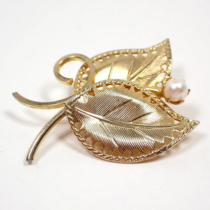 Front view of the Mid Century vintage leaves brooch pin. The metal is gold tone in color. There are two leaves with curved stems. They have a textured line pattern. There is a glass faux pearl at the top of the leaves.