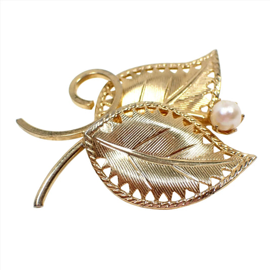 Front view of the Mid Century vintage leaves brooch pin. The metal is gold tone in color. There are two leaves with curved stems. They have a textured line pattern. There is a glass faux pearl at the top of the leaves.