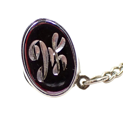 Enlarged front view of the Mid Century vintage initial tie tack. It is oval shaped and has a dark metallic gray faux hematite front. There is a fancy script letter H engraved in the middle. The rest of the metal is silver tone in color.