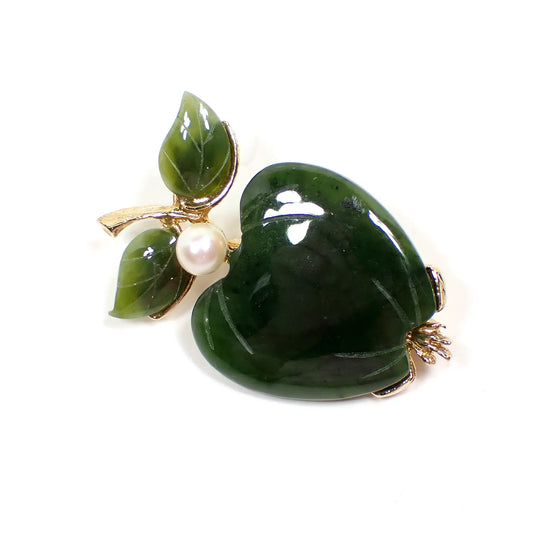 Angled view of the Mid Century Vintage Swoboda brooch pin. It is shaped like an apple with carved green jade apple shape and leaves by the stem. The metal is gold tone in color. There is  a cultured pearl at the top of the apple.