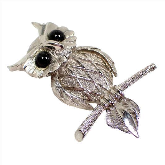 Angled view of the retro vintage owl brooch pin. It is silver tone in color and shaped like an owl sitting on a branch. There is a textured matte diamond pattern design on it's chest and it has black plastic eyes.