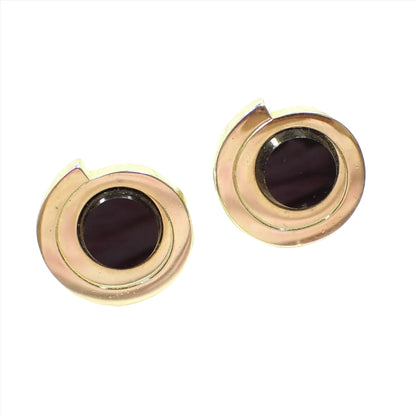 Angled front view of the retro vintage Pierre Cardin cufflinks. The metal is gold tone in color. They look like a bar that is shaped around itself in a circle shape. There are round flat top black onyx gemstone cabs in the middle.