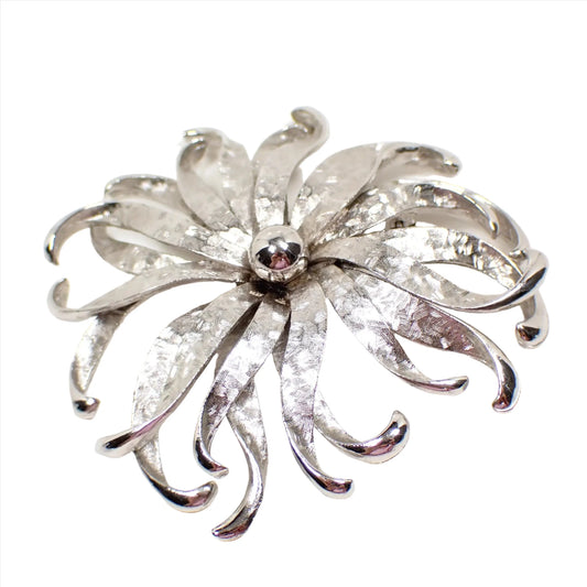 Front view of the retro vintage large flower brooch pin. It is silver tone metal with a brushed matte design. It is shaped like a flower with thin curvy petals all the way around.