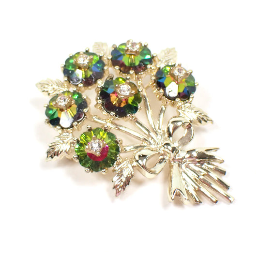 Angled view of the Mid Century vintage Emmons rhinestone brooch pin. The metal is gold tone in color. It is shaped like a bunch of flowers tied with a ribbon bow. The flowers are watermelon color flower shaped rhinestones with green, yellow, and pink. In the middle of each flower is a small clear rhinestone.
