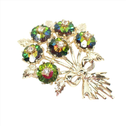 Angled view of the Mid Century vintage Emmons rhinestone brooch pin. The metal is gold tone in color. It is shaped like a bunch of flowers tied with a ribbon bow. The flowers are watermelon color flower shaped rhinestones with green, yellow, and pink. In the middle of each flower is a small clear rhinestone.