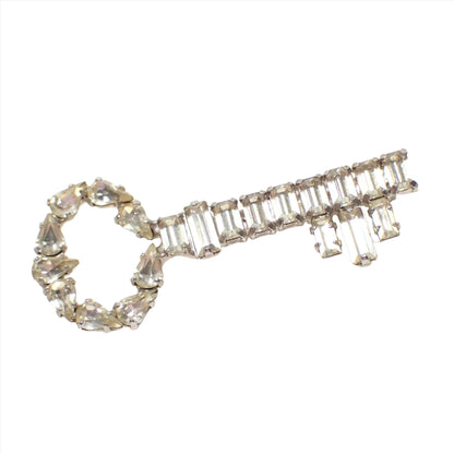 Angled view of the Mid Century vintage B David brooch pin. It is shaped like a skeleton key with silver tone color metal. There are clear teardrop shaped rhinestones around the top part of the key and baguette shaped rhinestones on the rest. The rhinestones are slightly yellowed from age.