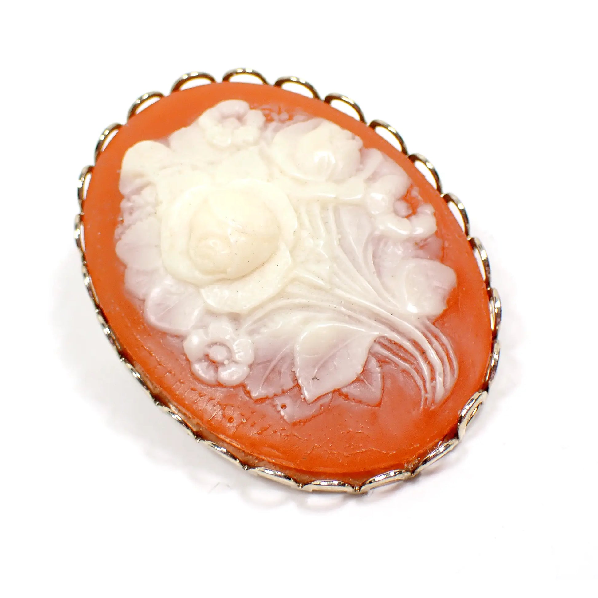Angled front photo of the vintage molded plastic floral cameo brooch. It has an oval shape with flowers in the middle in an off white light yellow color. The background is salmon orange and the setting is gold tone plated.