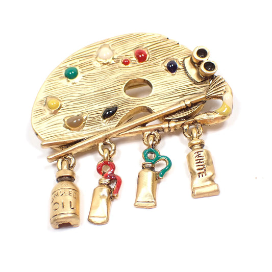 Front view of the Danecraft vintage brooch pin. It is shaped like an artist's palette with two paintbrushes across it. Dangling from the bottom are four charms. Three are paint tubes for white, green, and red. The last says Linseed oil. There is colored enamel on the red and green charms and on the palette in multiple colors.