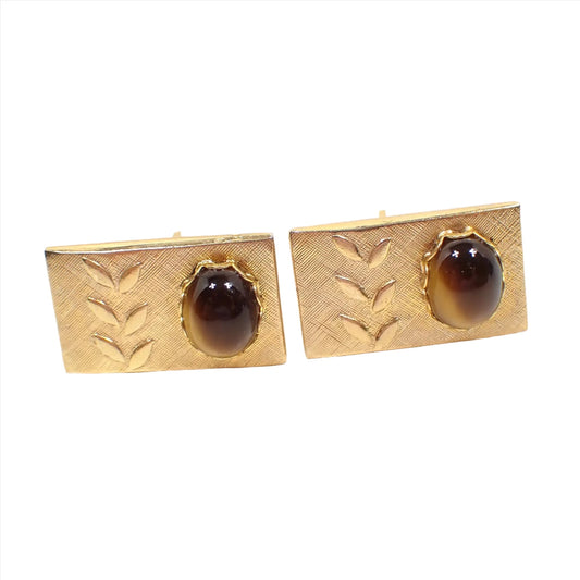 Front view of the Mid Century gemstone cufflinks. The cuff links are rectangle shaped with gold tone plated metal. There is a raised leaf design on one side and domed oval tiger's eye cabs on the other.