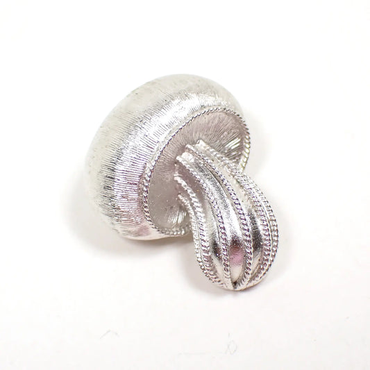 Front view of the retro vintage Crown Trifari brooch pin. The metal is matte silver tone in color and textured with lines. It is shaped like a small mushroom with a curved stem.'