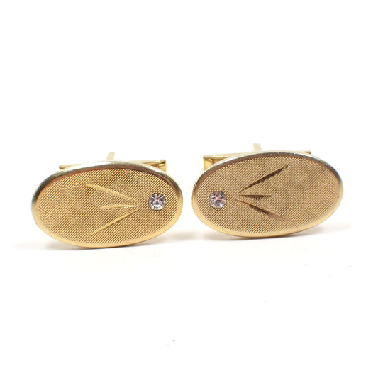 Front view of the Mid Century vintage cufflinks. The metal is gold tone in color. They are oval shaped and have brushed matte fronts. There is a small clear rhinestone on one side and diamond etched angled lines in the middle.