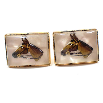Angled front view of the Mid Century vintage Anson cufflinks. They are rectangular in shape with a gold tone color setting. The fronts have clear crystal glass with a horses head carved and painted in reverse to give it a semi 3D like appearance.