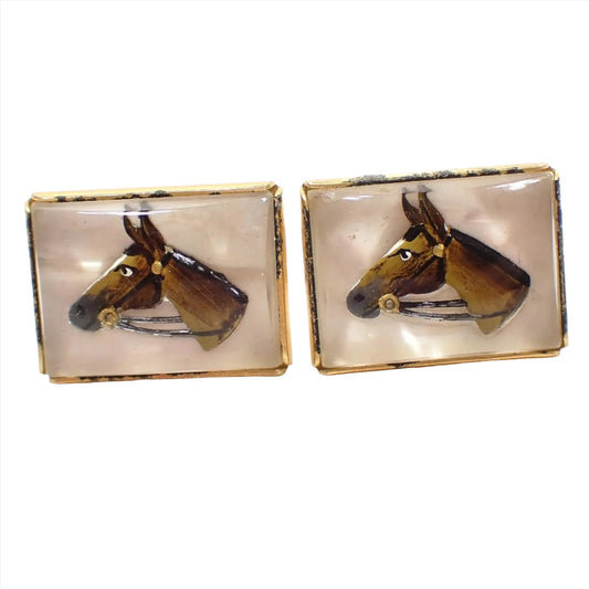 Angled front view of the Mid Century vintage Anson cufflinks. They are rectangular in shape with a gold tone color setting. The fronts have clear crystal glass with a horses head carved and painted in reverse to give it a semi 3D like appearance.