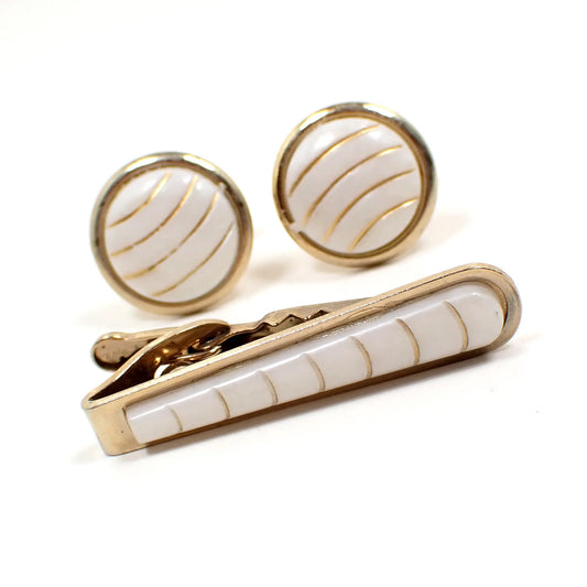 Angled view of the retro vintage tie clip and cufflinks men's jewelry set. The cufflinks are round with white plastic cabs on the front that have curved gold color painted stripes. The tie clip has a rounded end and has a matching plastic cab on the front.