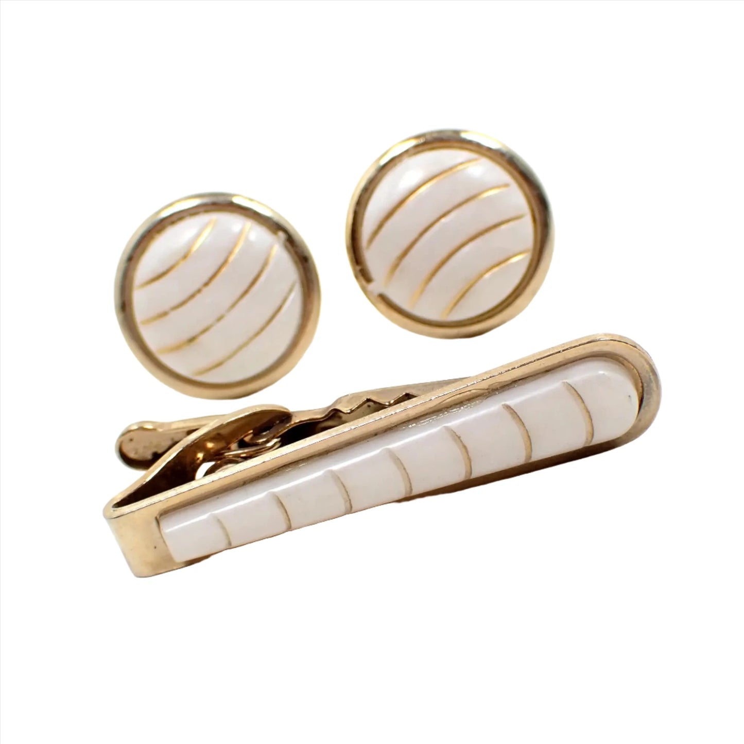 Angled view of the retro vintage tie clip and cufflinks men's jewelry set. The cufflinks are round with white plastic cabs on the front that have curved gold color painted stripes. The tie clip has a rounded end and has a matching plastic cab on the front.