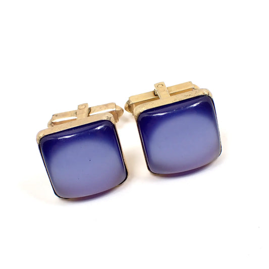 Angled view of the Mid Century vintage Anson moonglow lucite cufflinks. They have a square shape with rounded edges and a gold tone metal color setting. The front cabs are blue in color and have a glow like effect as you move around in the light.