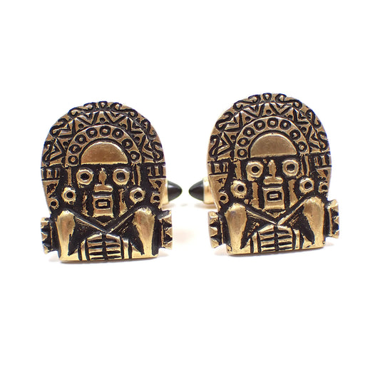 Front view of the Mid Century vintage Krementz cufflinks. The metal is antiqued gold tone in color with black accents. The fronts have a Aztec Mayan Sun God design. Part of the bullet backs are showing in the photo with pointed domed black lucite ends.