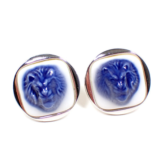 Front view of the Swank and Royal Copenhagen vintage cufflinks. The cufflinks are round with silver tone color metal. The porcelain cabs are rounded square shaped and are white with a blue roaring lion's head protruding from the middle. 