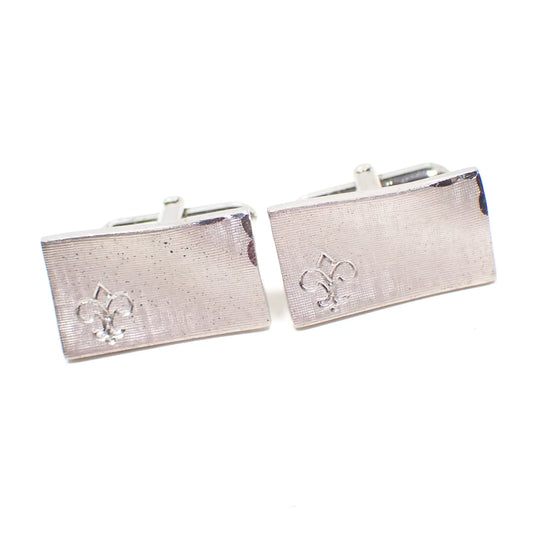 Angled front view of the Mid Century vintage Swank cufflinks. The metal is silver tone in color and the fronts have a brushed matte appearance. There is an etched Fleur de Lis design on the bottom left corner and and etched edge design on the top right corner.